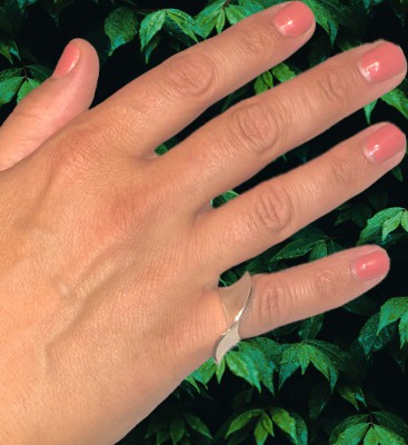 A silver mobius shaped pinky ring on a hand that has pink nail polish and is in front of a plant