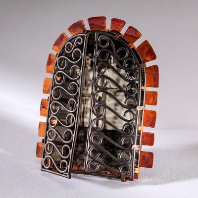 angled view of a brooch with copper rays, silver wire gates in the shape of wrought iron that are open with a black and white photo behind the partially open gates