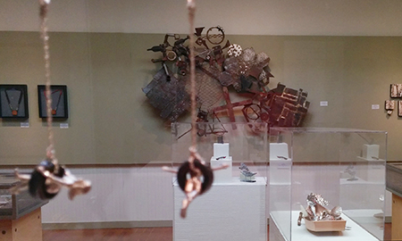 Horizontal photo of the inside of the Pacific Art League gallery with the installed Small Works show. On the wall ahead is a large metal sculpture with many different textures and shapes and a void in the center. Close to the viewer, and out of focus, are earrings with blacked silver tire swing shapes and human shapes swinging on those tires. In the lower right is a handmade wooden boat as seen in some sushi restaurants. In the boat are a variety of mokume gane shell shapes and kelps