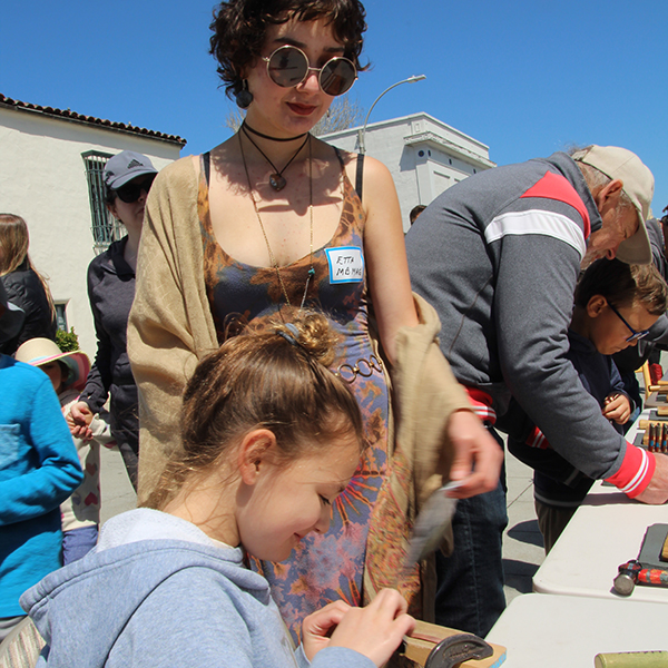 A new guild member volunteer helps as a participant sands a copper strip to create a bracelet at the Make A Bracelet event as part of the Monterey Museum of Art Block Party 2023