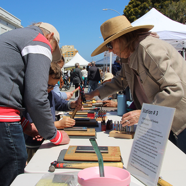 A young participant uses a hammer on a metal stamp to create a design on a copper strip to create a bracelet at the Make A Bracelet event as part of the Monterey Museum of Art Block Party 2023