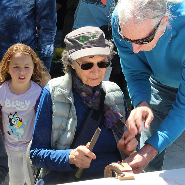 A guild metal artist volunteer teaches how to file a copper strip to create a bracelet at the Make A Bracelet event as part of the Monterey Museum of Art Block Party 2023