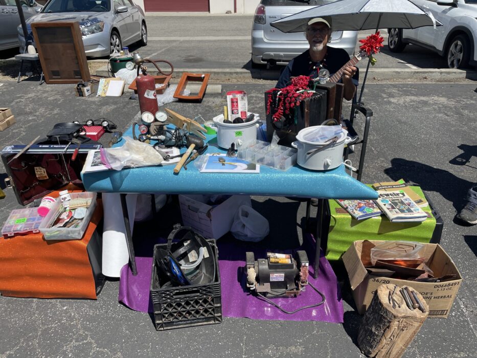 Jeff representing SMAC, entertaining while selling tools and supplies including a bench polisher, a stump, hand tools, optivisor, books, pickle pots,a tank and other items on a table and on the asphalt of the parking lot.