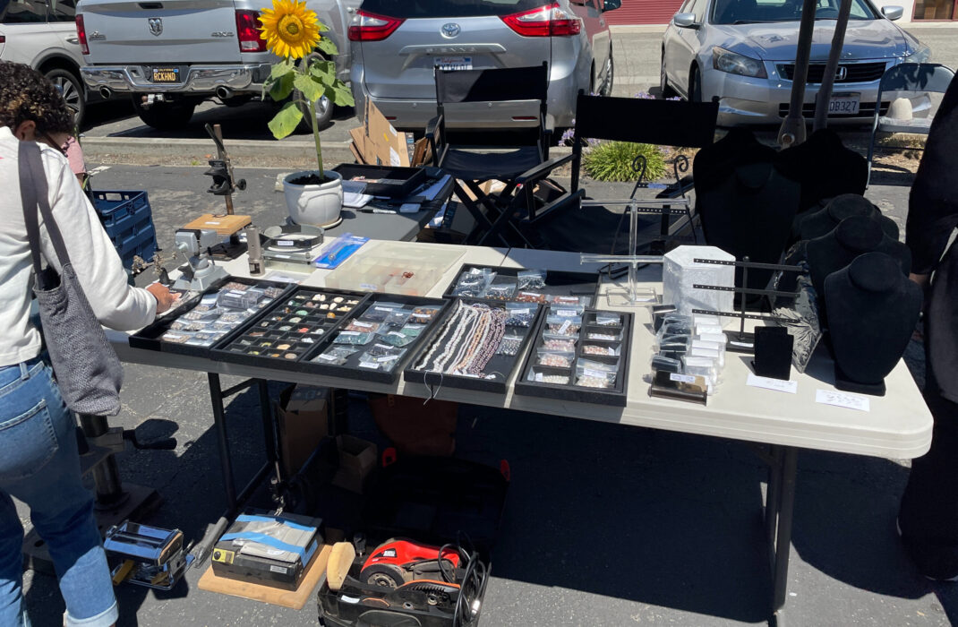 N Kay Morrison's setup for selling tools, stones, beads, bench pin, display items and more on a table with a sunflower plant and directors chairs.