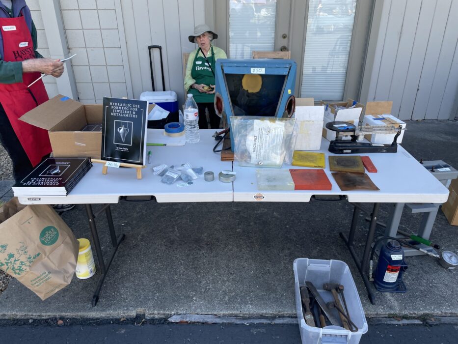 Susan Kingsley with copies of her books for sale and metalsmithing tools for sale: a hydraulic press, a scale, a sandblast cabinet with media, accessories for the hydraulic press, hammers and more.