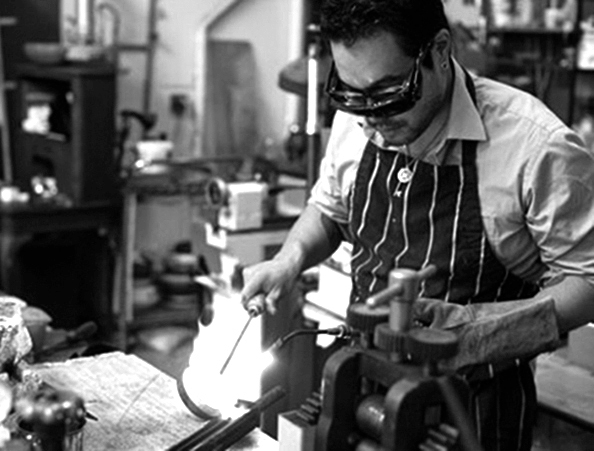 Black and white portrait of Curtis Arima melting metal in a crucible in his workshop. He is wearing eye protection and a fire resistant glove in his left hand. His apron has vertical stripes. He is wearing a necklace. In the foreground is the top of a rolling mill.