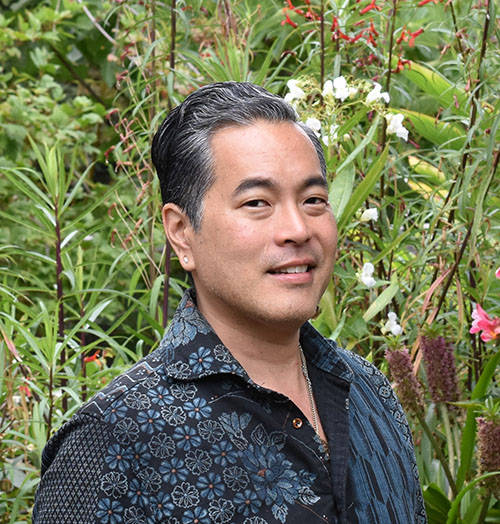Curtis Hidemasa Aria Portrait Headshot. Curtis is wearing a dark hand made shirt standing in front of greenery in a garden.
