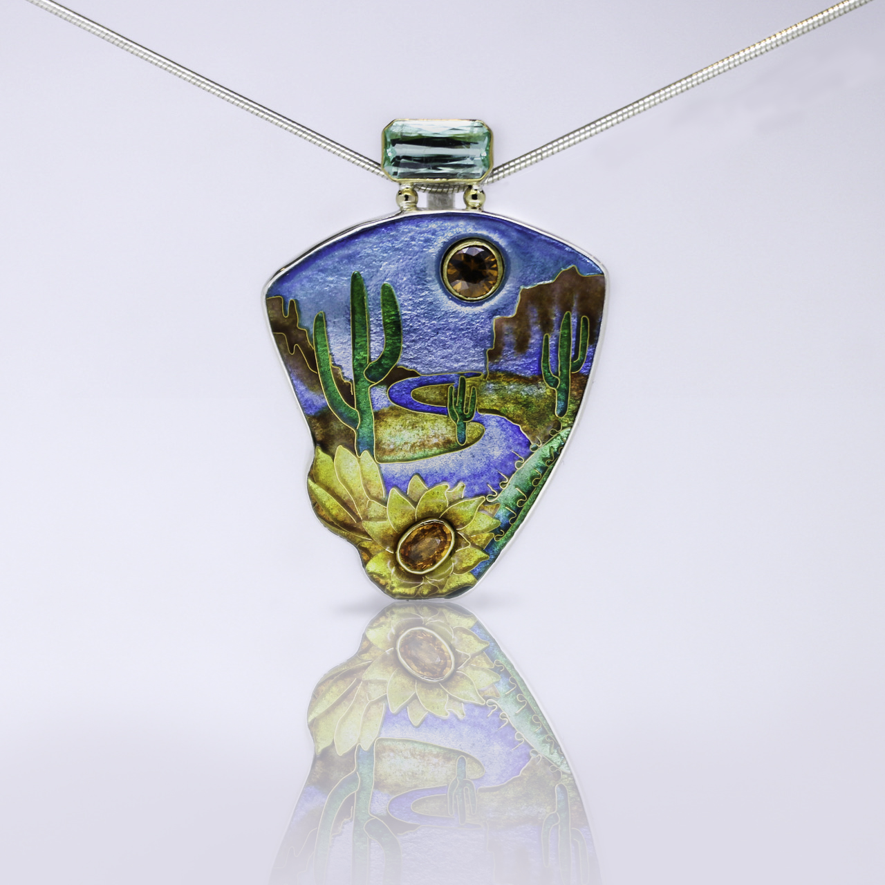 Landscape cloisonné enamel pendant by Darci Shea Bogdan is a scene in a canyon featuring cactus along a meandering river, a flower with a faceted yellow stone center, and a yellow faceted stone sun set into the sky